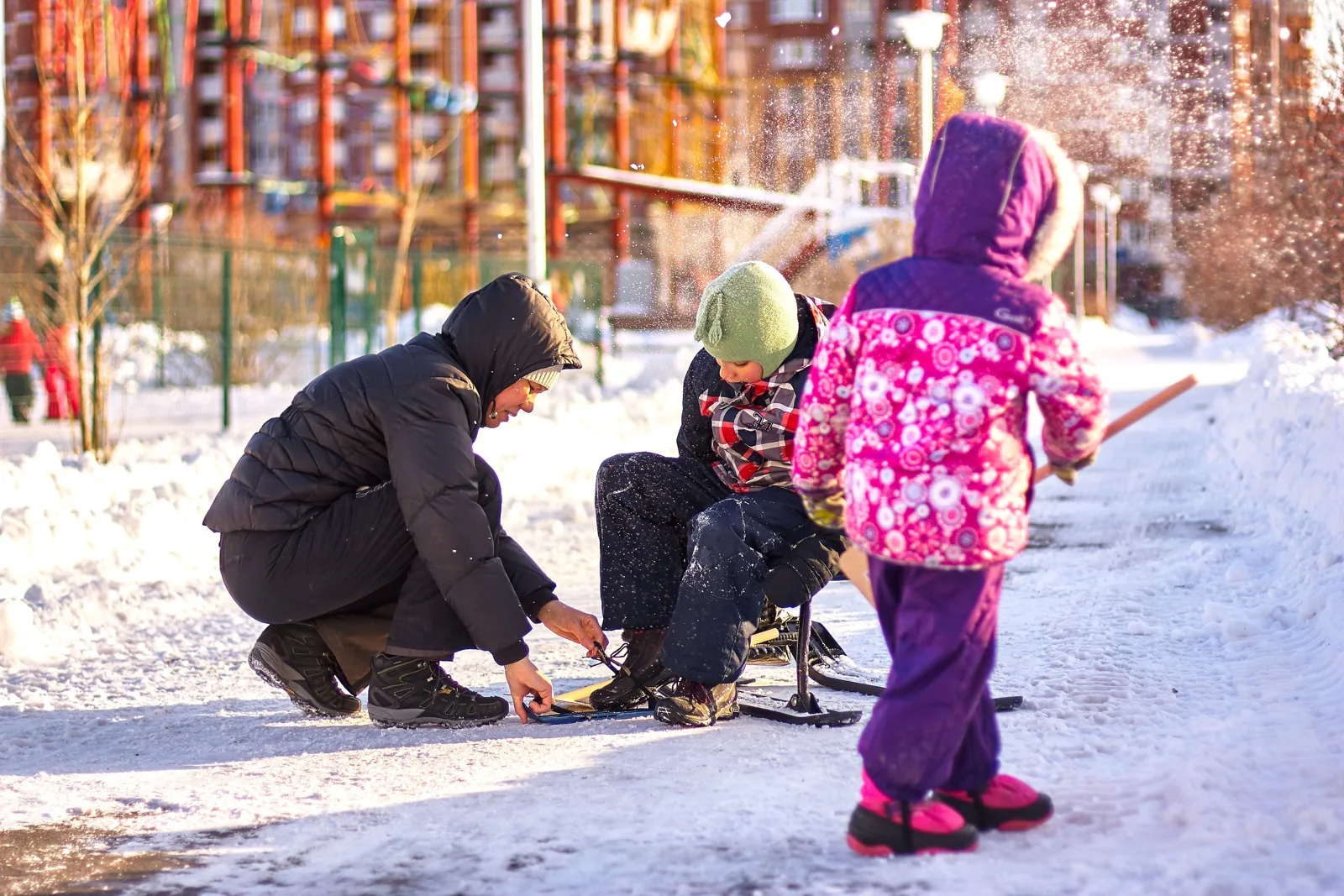 Un père charge des skis près du coffre ouvert de la voiture, la famille l’attend dans la neige avec une luge